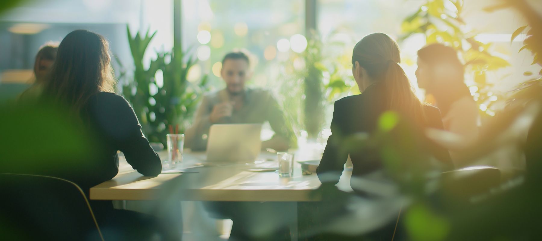 Gruppe von Personen in einem modernen Büro bei einer Besprechung, umgeben von großen Fenstern und grünen Pflanzen