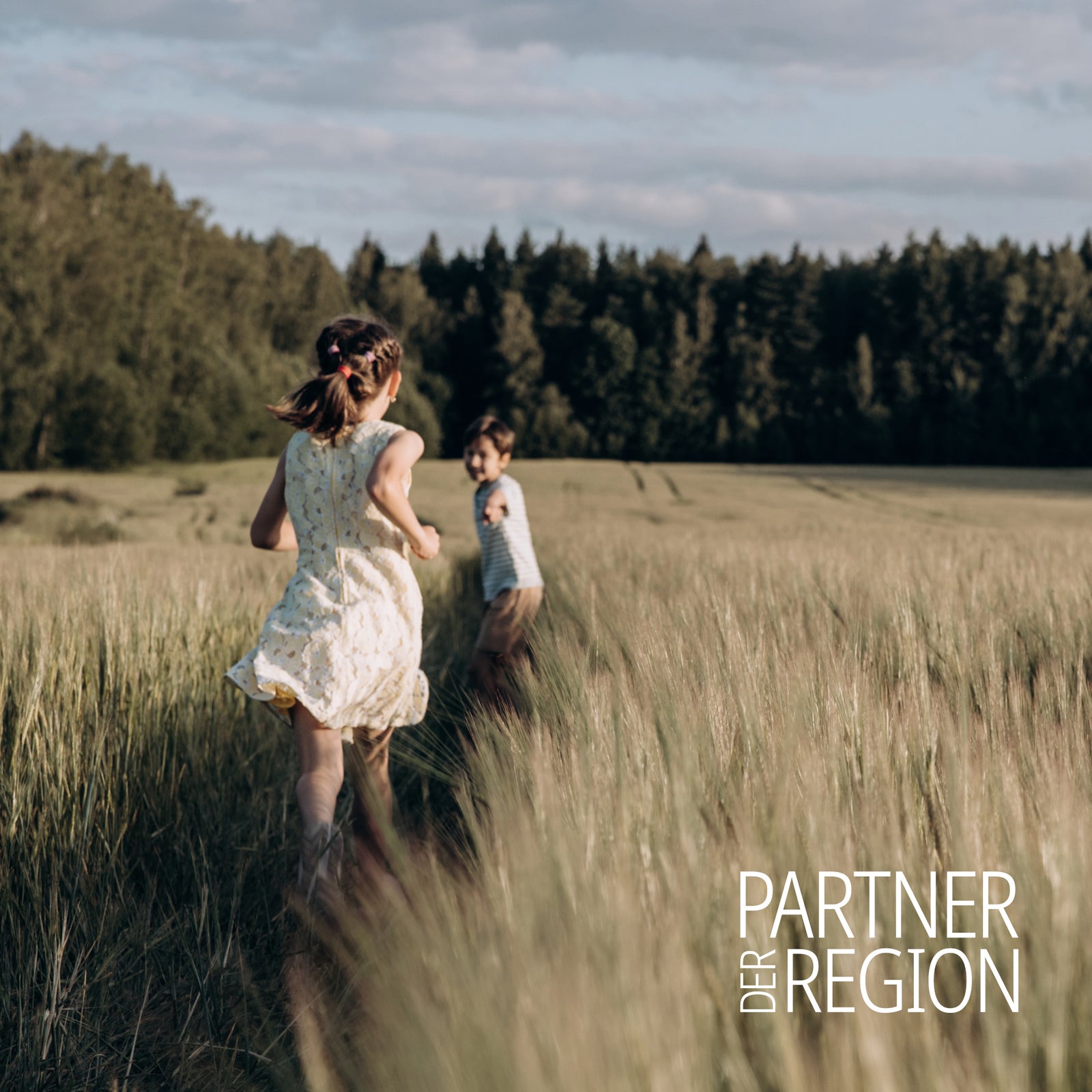 Zwei Kinder spielen fröhlich in einem weiten Getreidefeld vor einem Waldhintergrund. Logo von "Partner der Region" ist unten rechts in der Ecke erkennbar.