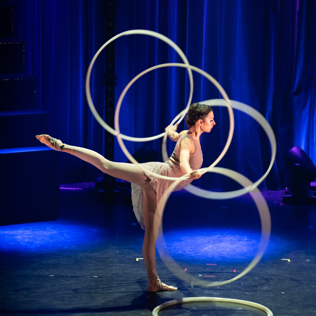Eine Person führt eine elegante Hula-Hoop‑Nummer auf der Bühne aus und balanciert mehrere große Reifen gleichzeitig, während ein Bein seitlich ausgestreckt ist
