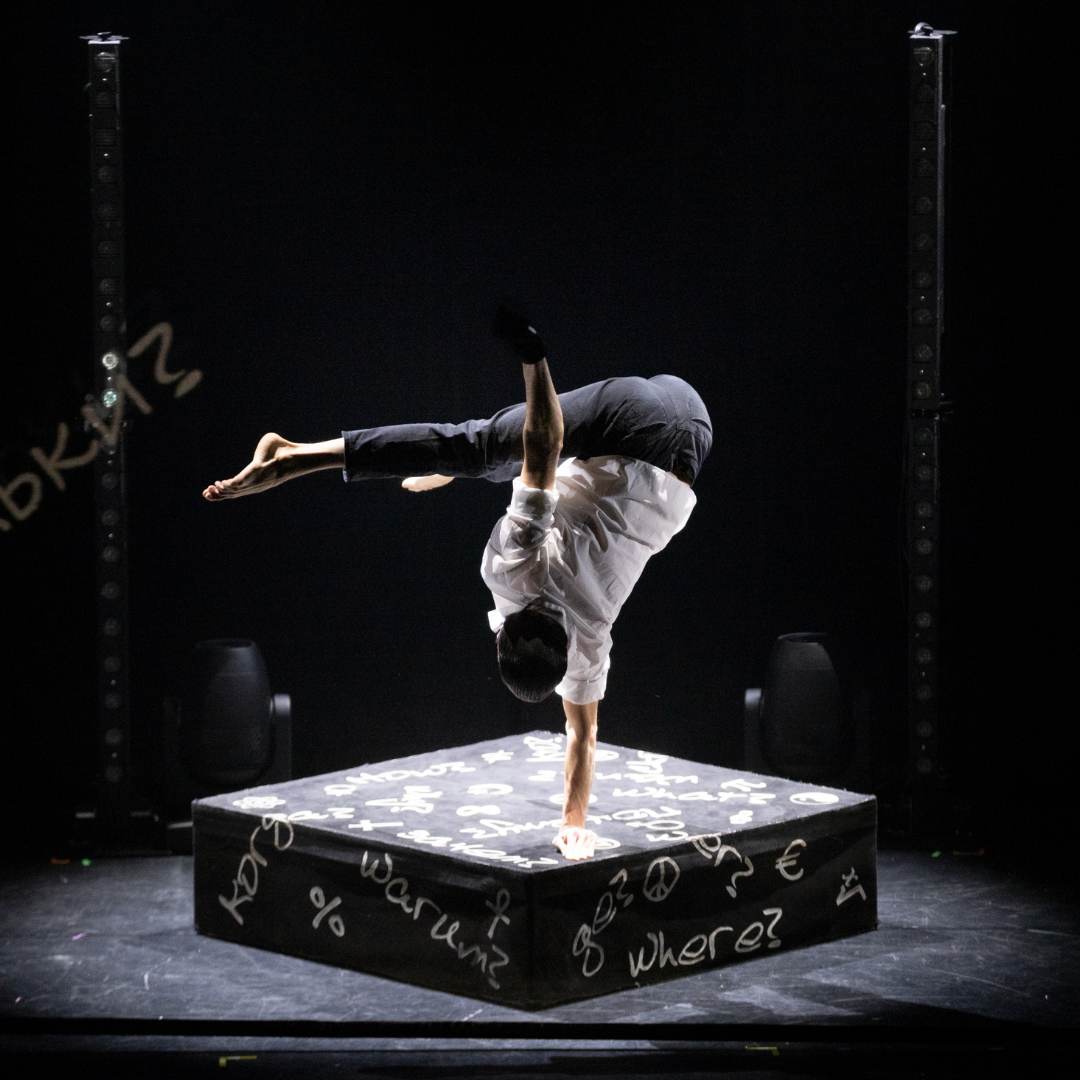 Eine Person führt auf einer erhöhten Plattform eine einarmige Balancefigur aus, während die Beine dynamisch nach oben geschwungen sind und der Bühnenhintergrund dunkel bleibt.