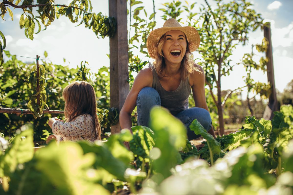 Eine lachende Frau und ein Kind inmitten von grünen Pflanzen, die in einem Garten arbeiten, symbolisieren Freude an der Natur und nachhaltige Landwirtschaft.