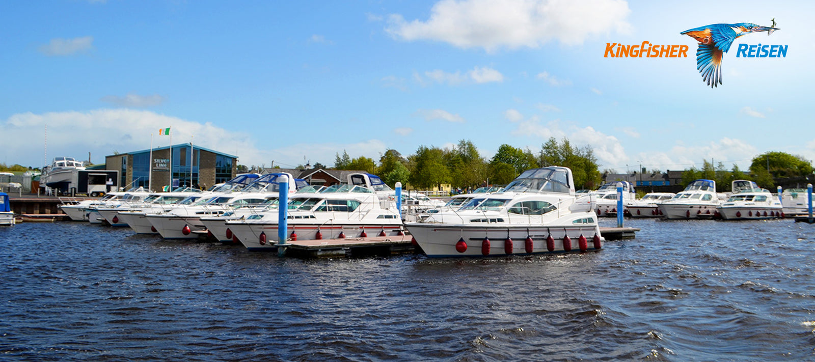  Eine Reihe von Freizeitbooten, die an einem sonnigen Tag an Docks in einem Hafen vertäut sind, mit einem modernen Gebäude im Hintergrund. 