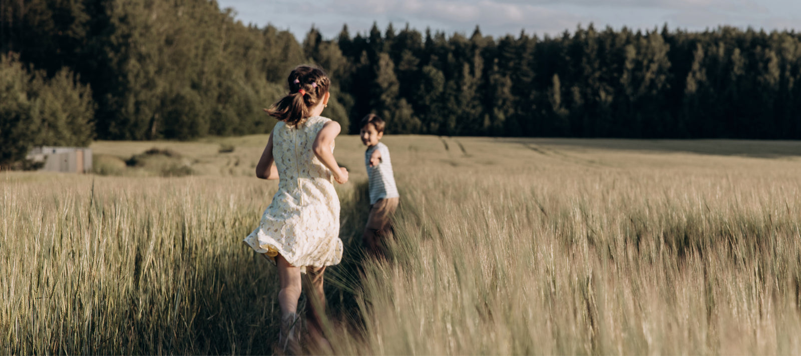 Zwei Kinder spielen fröhlich in einem weiten Getreidefeld vor einem Waldhintergrund.