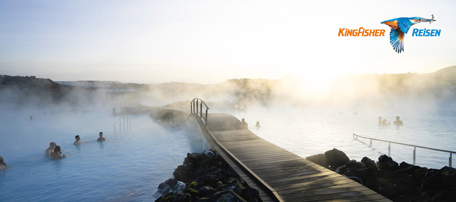  Panoramaaufnahme eines geothermalen Bades, umgeben von Felsen, mit Menschen, die im dampfenden blauen Wasser entspannen. Ein hölzerner Steg führt durch den Nebel. 